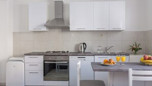 Una cocina moderna con armarios blancos, horno integrado, lavavajillas y cafetera. Hay una mesa de comedor con sillas y un cuenco de fruta.