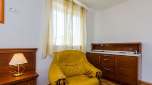 Ein gemütlicher Raum mit Holzstuhl, einem kleinen Tisch mit Lampe, einer Küchenzeile mit Spüle und einem Fenster mit Vorhängen.