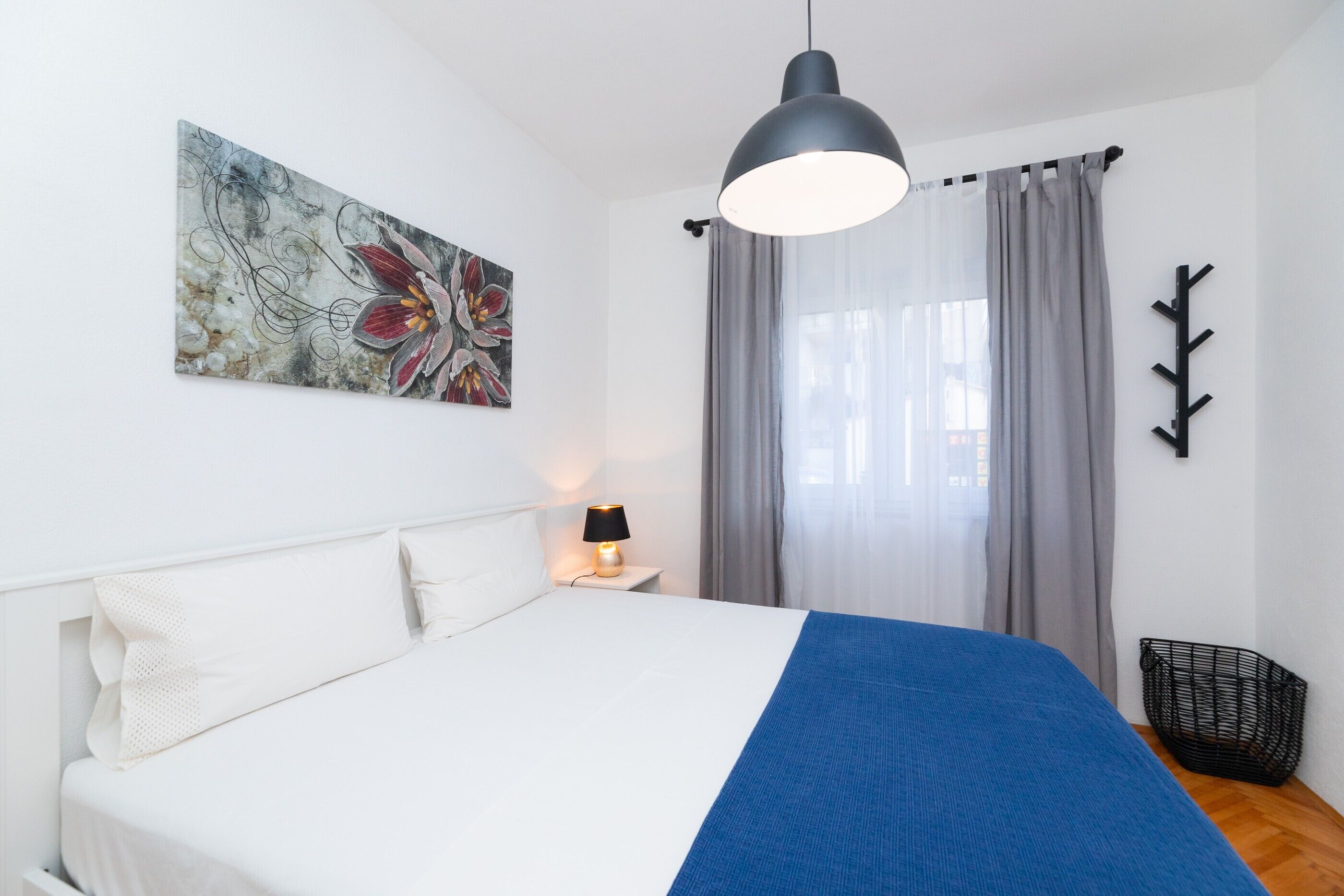 A bedroom with a bed, a bedside lamp, a wall-mounted artwork, a window with curtains, and a black coat rack.