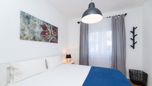 Een slaapkamer met een bed, een bedlampje, een kunstwerk aan de muur, een raam met gordijnen en een zwarte kapstok.