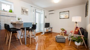 Uma sala de estar moderna com piso de madeira, uma área de jantar com duas cadeiras, uma televisão e um sofá. Há uma janela com persianas, um abajur e um espelho na parede.