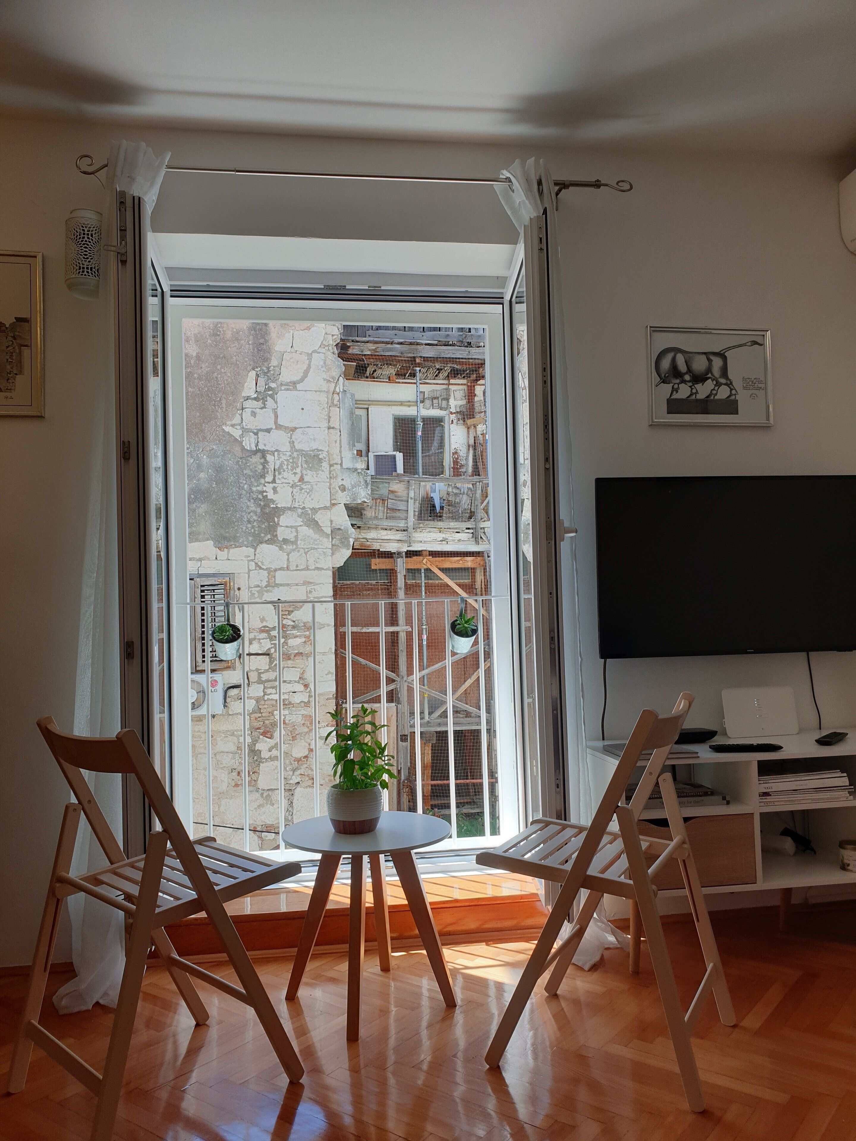 A living room with a large window, a flat-screen TV, a table with a plant, and two wooden chairs.