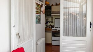 Ein Zimmer mit einem roten Sessel, einem Tisch mit weißer Spitzenleinentuch, einer weißen Tür mit Fenster und einer durch die Tür sichtbaren Küche.