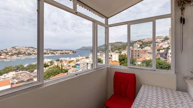 Ein Zimmer mit einem großen Fenster, das einen Blick auf eine Küstenstadt bietet, ein roter Sessel und ein Tisch mit Muster.