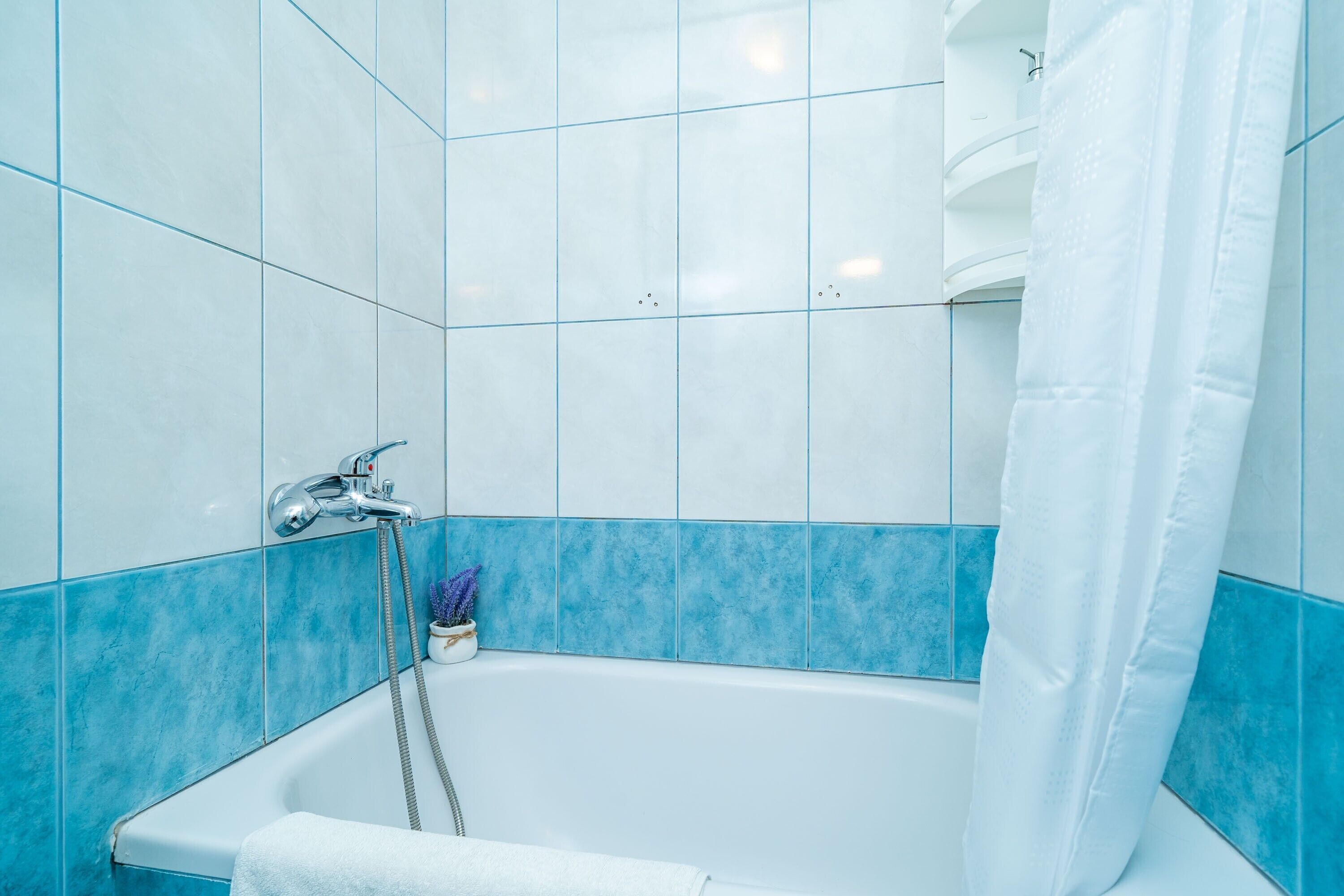 A bathroom with a white bathtub, blue tiles, a shower curtain, and a small vase with purple flowers.