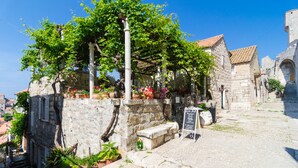 Ein Steinhaus mit einer Pergola, die in Grün bewachsen ist, mit Topfpflanzen und einem Kreidetafel-Schild.