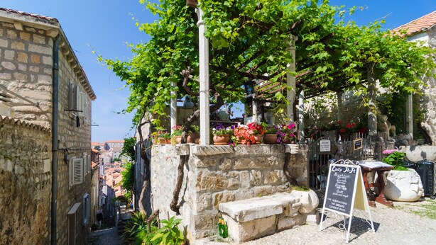 Eine enge Gasse mit Steinhäusern, einer Steinsitzbank und einem Schild für ein Café.