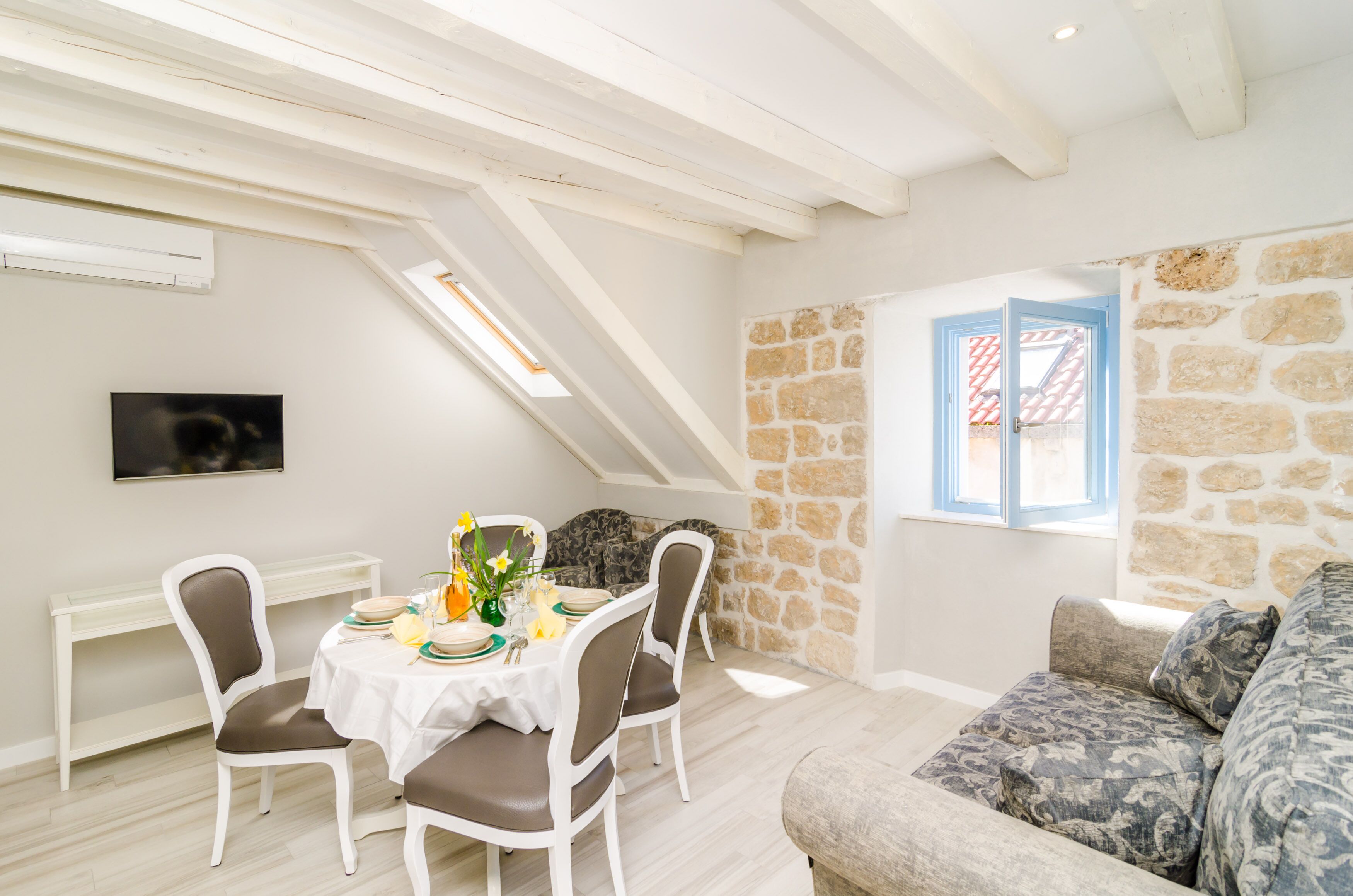 Un coin repas avec une table dressée pour quatre personnes, un mur en pierre et une fenêtre avec des volets bleus.