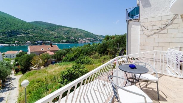 A balcony with a table and chairs overlooking a coastal area with houses and a mountain. - Apartments Peco - Four Bedroom Apartment with Balcony and Sea View (Mokosica)