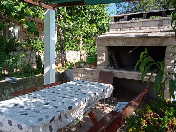 Enjoy al fresco dining under a shaded pergola surrounded by lush greenery and a stone fireplace.
