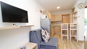 Un espace de vie compact comprenant un canapé, un téléviseur à écran plat, un réfrigérateur, un four à micro-ondes, des meubles de rangement en bois, un coin repas avec des chaises et une horloge murale.
