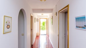 A hallway with red tiled flooring, white walls, and a wooden bench. There are framed pictures on the walls and a door on the right.