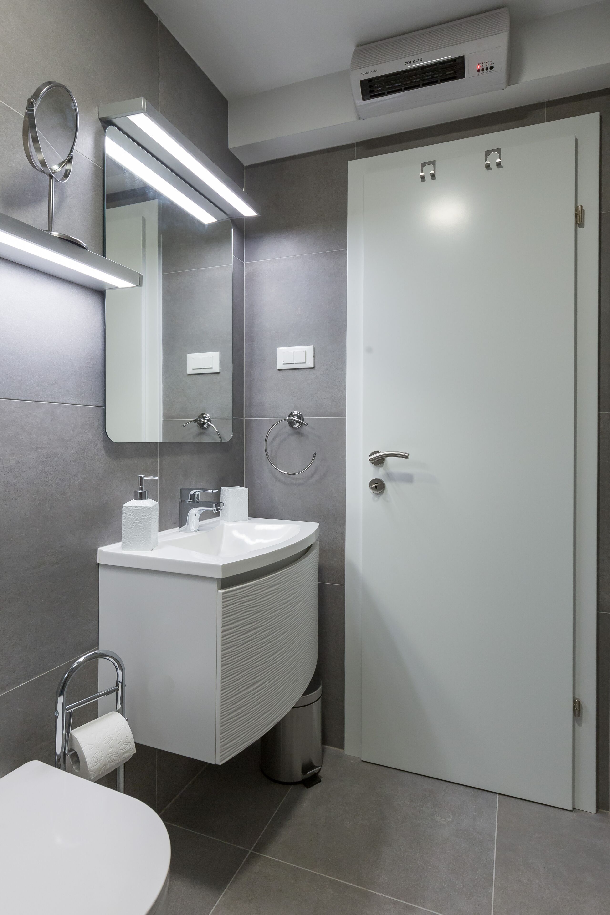 A modern bathroom with a white sink, mirror, and toilet. There is a towel rack, a trash bin, and a door with a handle.