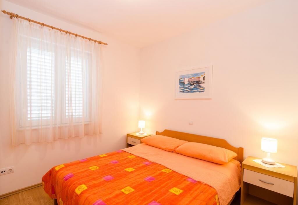 A bedroom with a bed, two bedside tables with lamps, a window with sheer curtains, and a framed picture on the wall.