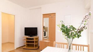 Ein Zimmer mit Fernseher auf einem Holztisch, einer kleinen Küchenzeile, einem Esstisch mit Blumen in einer Vase und einem Holzschrank.