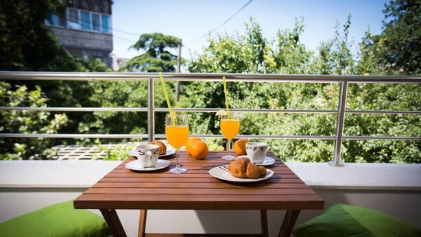 Une table en bois, dressée avec des aliments pour le petit-déjeuner, notamment des croissants, du jus d’orange et des tasses de café, est placée devant un balcon doté d’une rambarde en métal, qui offre une vue sur un espace verdoyant luxuriant.