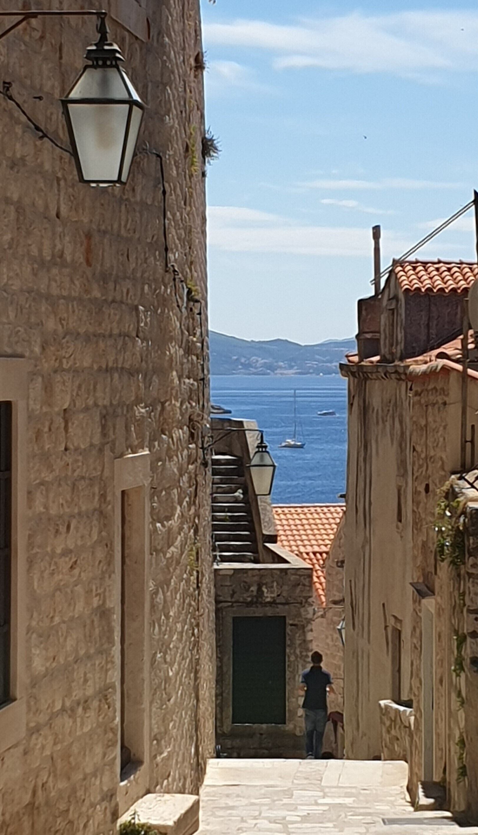 Une ruelle pavée étroite, où l’on aperçoit une personne qui marche, et qui mène à un point de vue sur la mer et les bateaux.