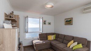 A modern living room with a sectional sofa, a wooden cabinet, a window with a view, and a ceiling light fixture. - Apartment Gigi - Two Bedroom Apartment with Terrace and Sea View (Dubrovnik)