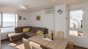 A modern living room with a wooden dining table, a brown sectional sofa, and a view of the sea through a large window. - Apartment Gigi - Two Bedroom Apartment with Terrace and Sea View (Dubrovnik)