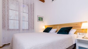A neatly made bed with a white bedspread and two pillows, a bedside table with a lamp, and a window with a sheer curtain.