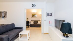 A modern living room with a grey sofa, a white coffee table, a TV, and a lamp. There is a door leading to another room with a couch and a lamp.