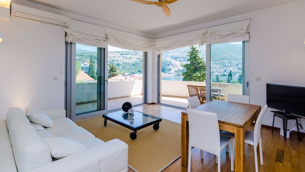 Ein modernes Wohnzimmer mit einer Couch, einem Glastisch, einem Esstisch mit Stühlen, einem Fernseher und einem Deckenventilator. Große Fenster bieten Blick auf die Stadt und die Hügel.