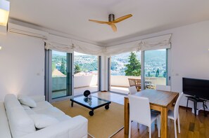 A modern living room with a sofa, a glass coffee table, a dining table with chairs, a TV, and a ceiling fan. Large windows offer a view of the city and hills.