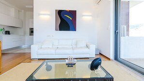 Ein modernes Wohnzimmer mit einem Glastisch, einem weißen Sofa und einem großen Fenster mit Blick ins Freie.