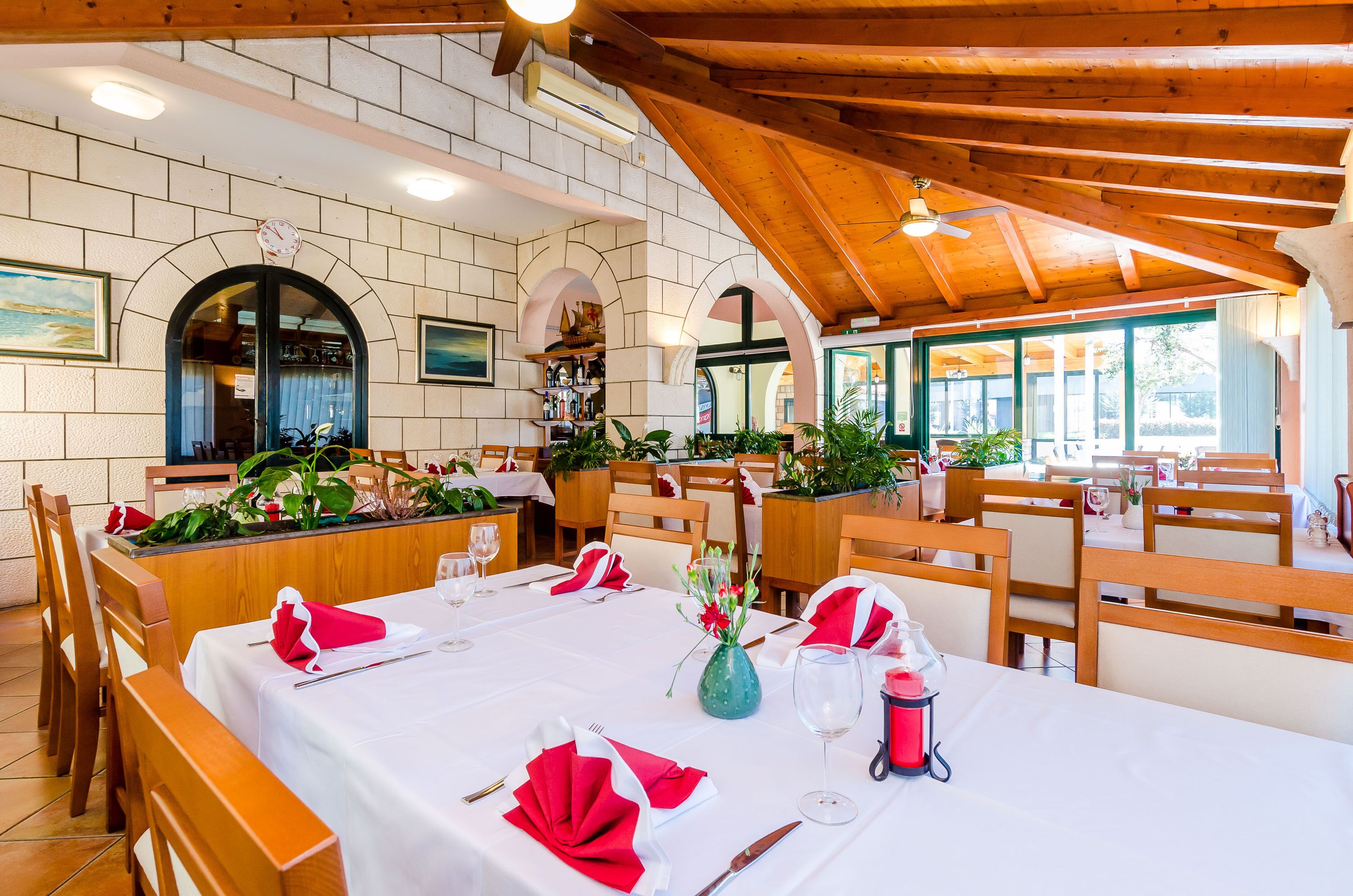 A restaurant with wooden beams, white walls, and arched doorways. Tables are set with red napkins and white tablecloths.