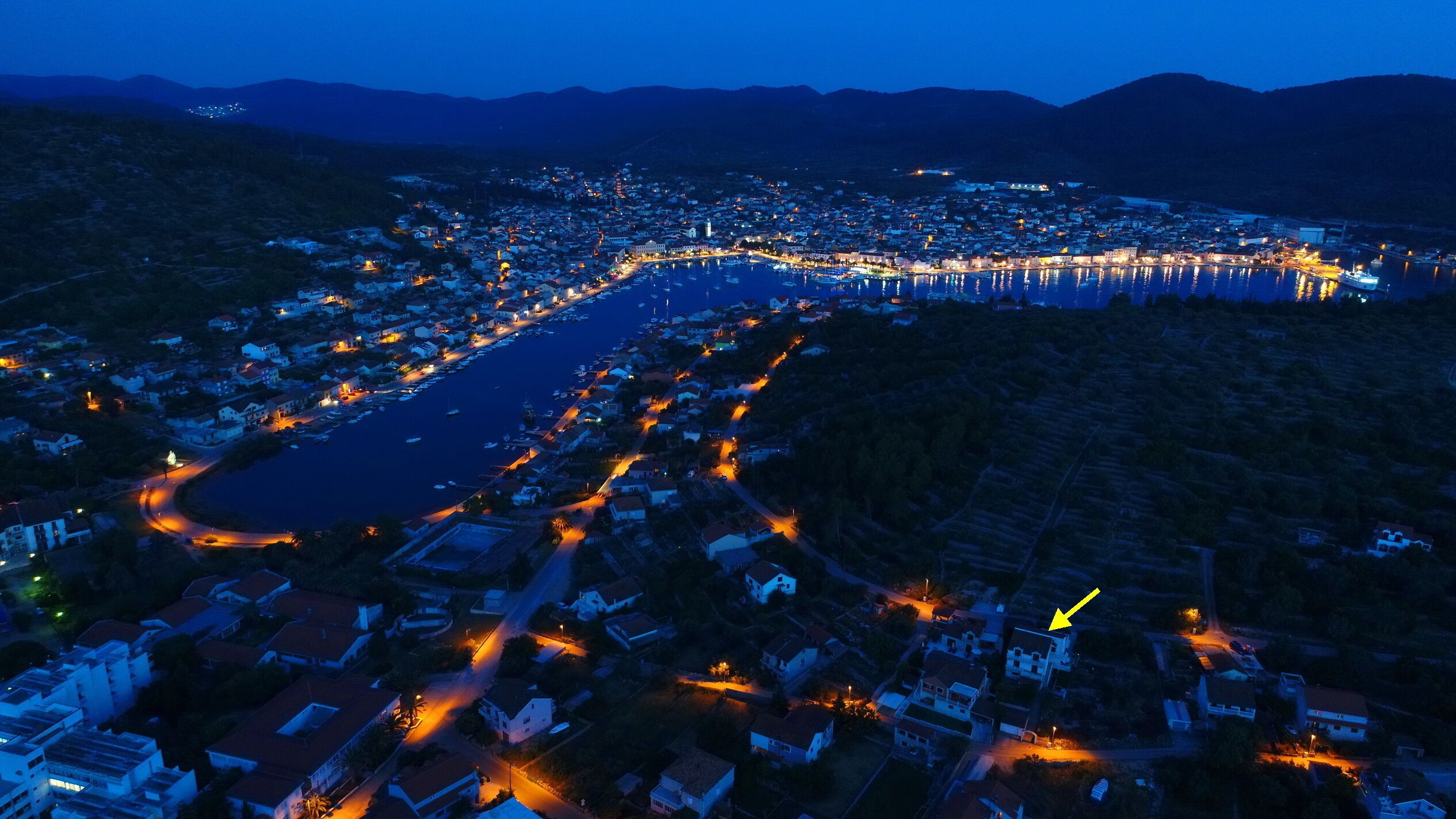 An aerial view of a coastal town at night with a marina, illuminated streets, and residential buildings.