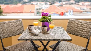 Un balcón con mobiliario de mimbre, dos tazas de café, un vaso de zumo de naranja y un jarrón con flores.