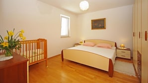Un dormitorio con una cuna de madera, una cama con ropa de cama blanca y rosa, una mesita de noche con una lámpara, un cuadro enmarcado en la pared y una ventana con persianas.