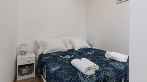 En snyggt bäddad säng med ett mönstrat överkast, ett vitt sängbord med en lampa och ett litet kassaskåp.