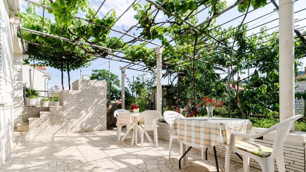 Een terras buiten met een tafel en stoelen, een bakstenen muur en een pergola begroeid met klimop.