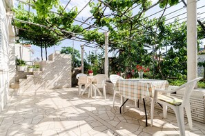 Een terras buiten met een tafel en stoelen, een bakstenen muur en een pergola begroeid met klimop.
