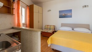 Quarto com cama, mesa de cabeceira, cadeira, quadro e área de cozinha com pia.
