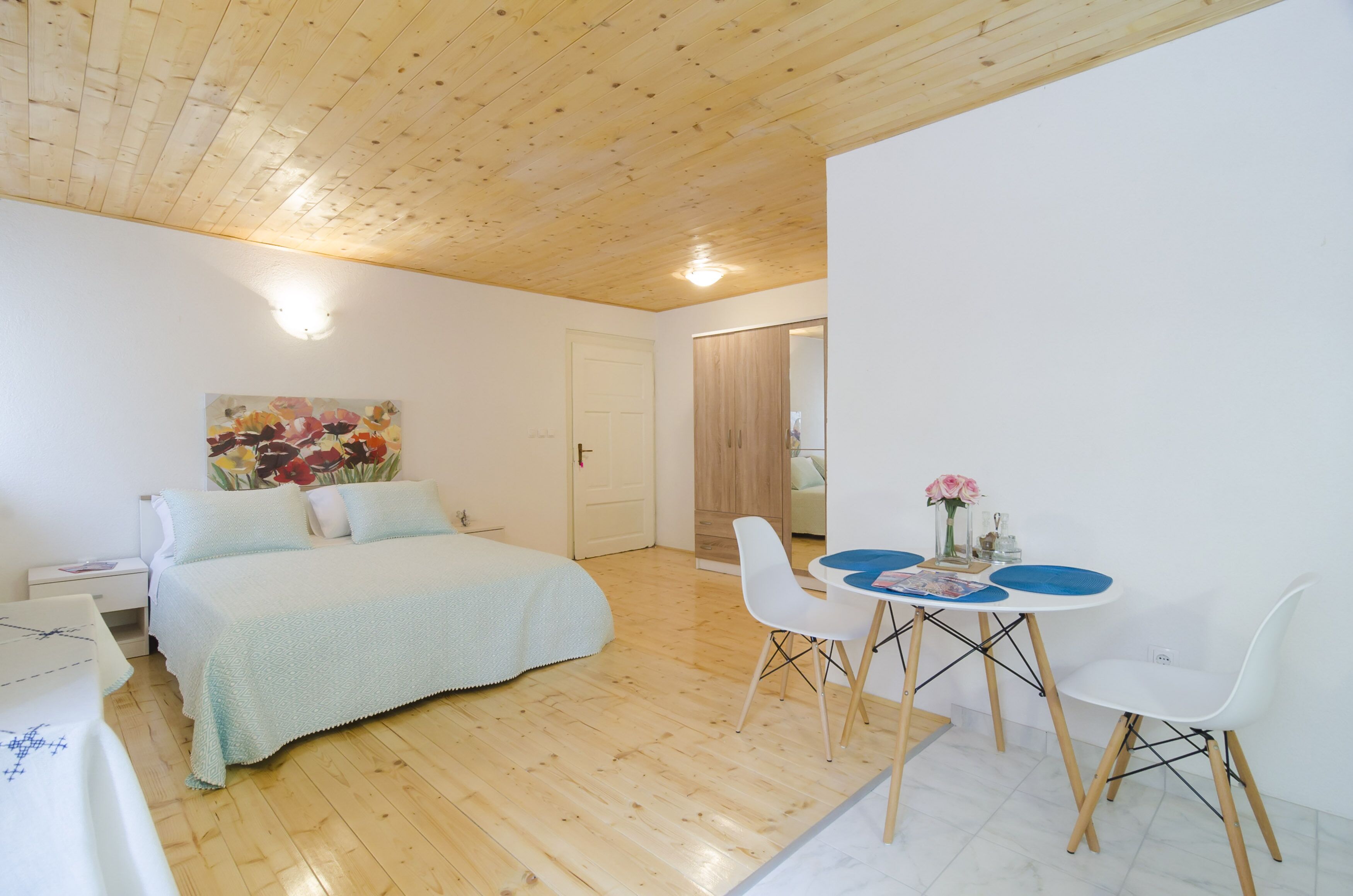 Un dormitorio con cama, mesita de noche, escritorio, silla, mesa con un florero y un cuadro en la pared.