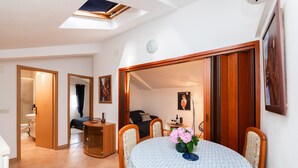 Une pièce comprenant une table à manger, des chaises, un luminaire au plafond et donnant sur une chambre à coucher par une porte ouverte.