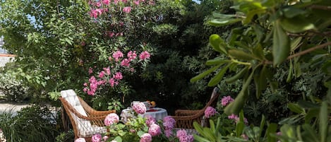 A garden with wicker furniture, a table with a bowl of fruit, and pink flowers.