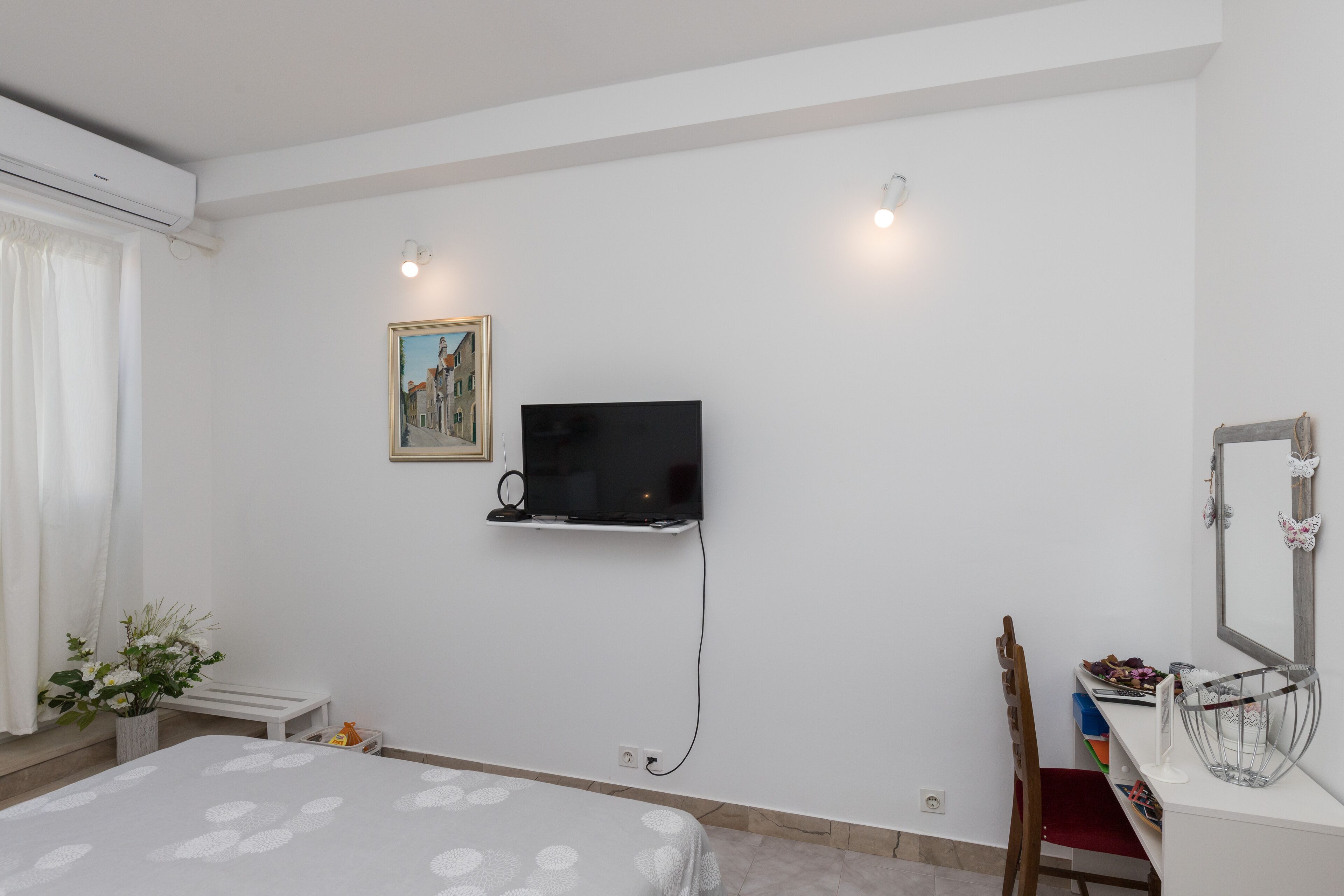 Une chambre à coucher avec un lit, un téléviseur fixé au mur, un bureau avec un miroir et une fenêtre avec des rideaux.