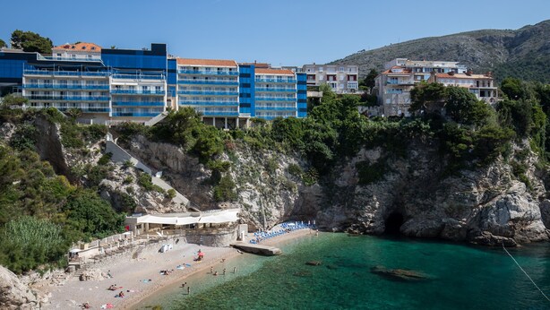A coastal hotel with multiple floors, a beach area with umbrellas, and a rocky cliff backdrop. - Room Primi - Double Room with Terrace (Dubrovnik)
