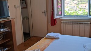 Une chambre à coucher avec un lit, une fenêtre donnant sur des maisons, un radiateur et un téléviseur posé sur un meuble.