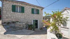 Un bâtiment en pierre avec deux fenêtres et une porte, entouré d’une zone pavée et d’un petit jardin.