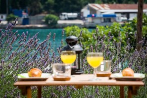 A table with two glasses of yellow drink, a lantern, and a plate with a fruit, set against a backdrop of purple flowers and a body of water. - Captain's Studio - Studio with Terrace (Mokosica)