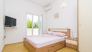 Une chambre avec un lit en bois, une table de chevet avec une lampe, un téléviseur fixé au mur et une porte-fenêtre coulissante donnant sur un balcon.