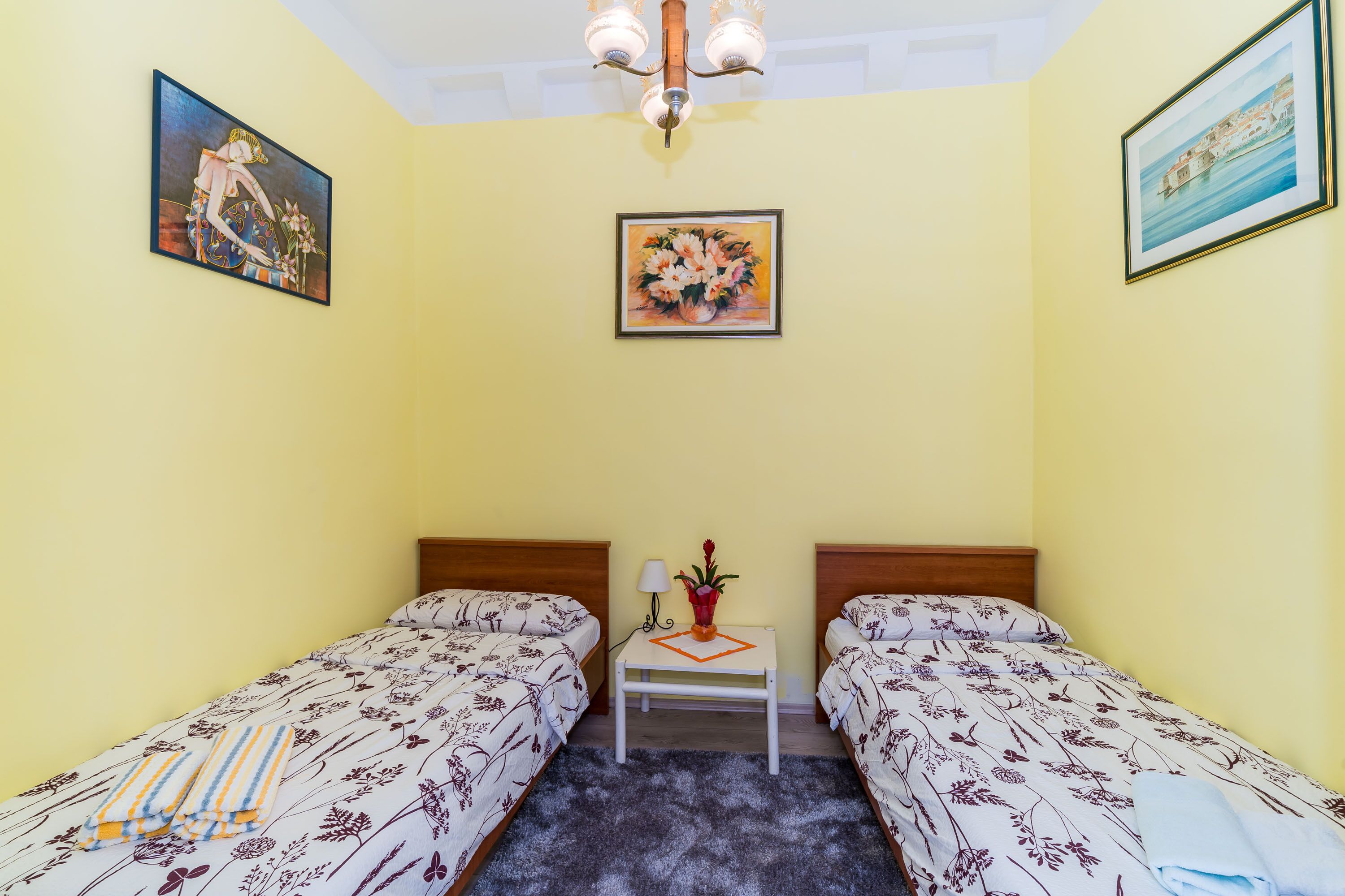 Ein Zimmer mit zwei Einzelbetten, einem kleinen Tisch mit einer Vase und zwei Bildern an den Wänden.