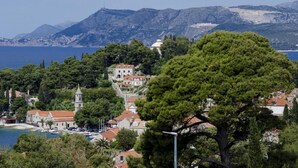 Eine Küstenstadt mit einer markanten Kirche, einem Gebäude auf einem Hügel und freiem Blick auf das Meer und die Berge im Hintergrund.
