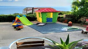 Eine Terrasse mit Spielbereich, inklusive Rutsche und einem kleinen Spielhäuschen, mit Blick auf ein Gewässer.