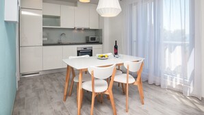 Une cuisine moderne avec des meubles blancs, une table à manger entourée de quatre chaises et une grande fenêtre ornée de rideaux légers.
