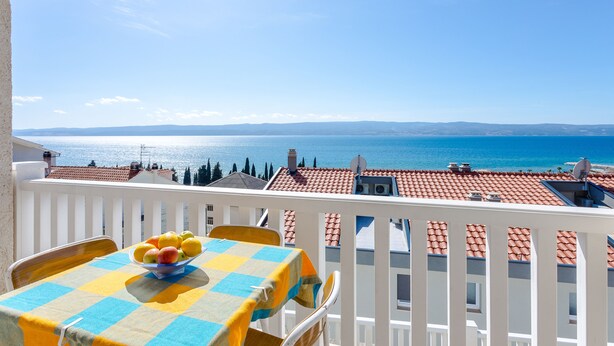 A balcony with a table and chairs, a bowl of fruit, and a view of the sea and distant mountains. - Magdales - Comfort Three Bedroom Apartment (Omis)